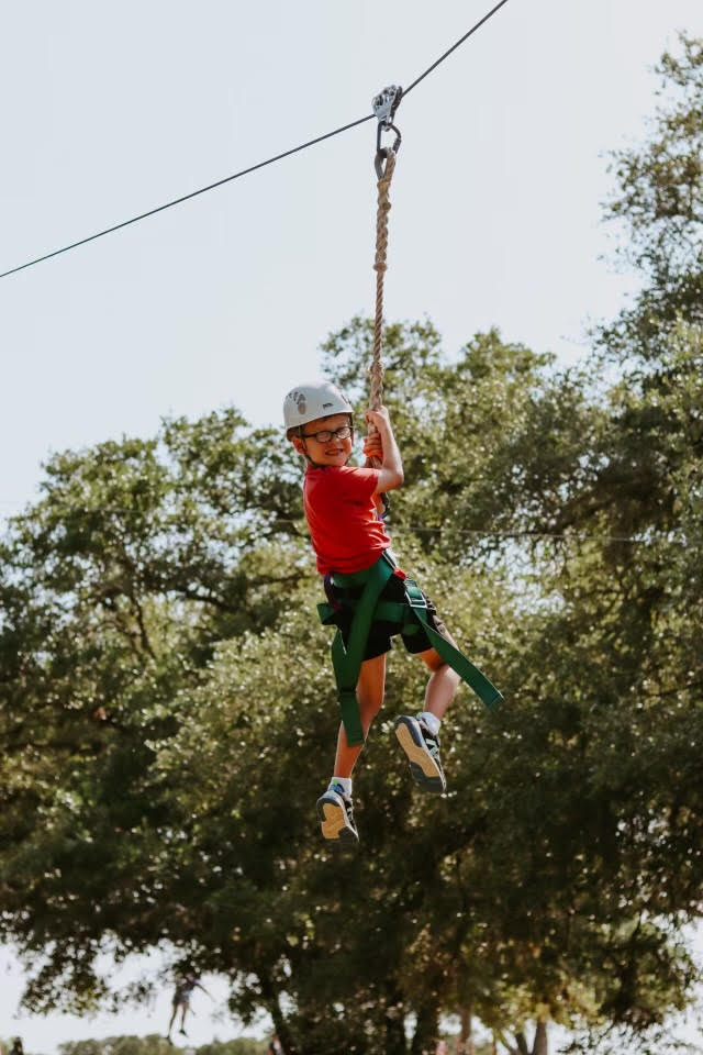 unnamed Young boy wearing a helmet, smiling as he holds onto a harness and wire on a zip line above the ground